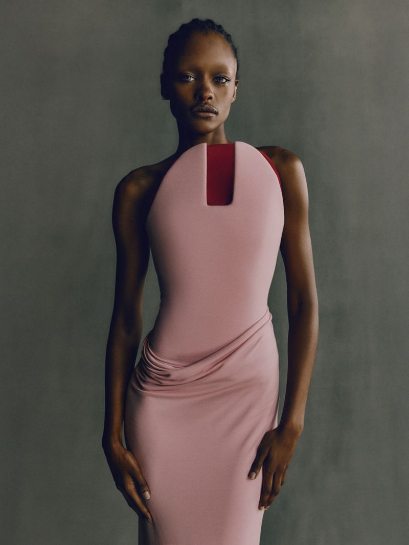 Fashionable African woman in a light pink dress posing in a studio. The dress features a unique square neckline and draped skirt.