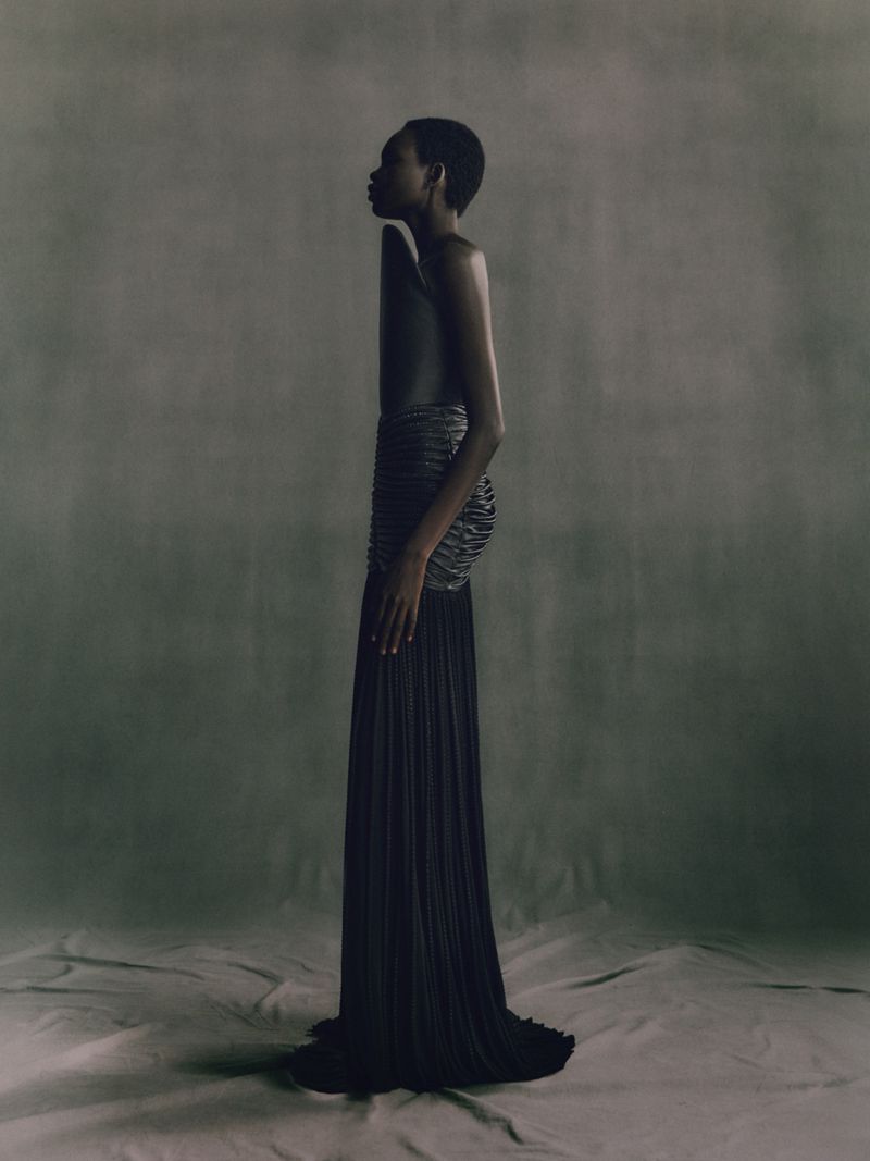 Striking studio portrait of an adult woman in an elegant black dress with intricate details, side profile, on a neutral-toned backdrop.