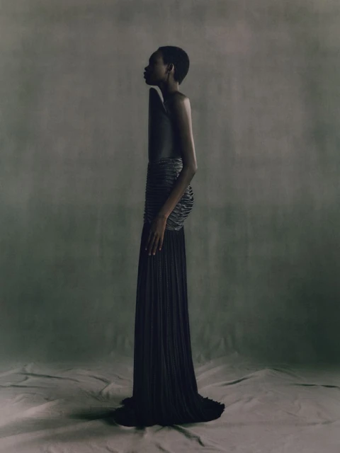 Striking studio portrait of an adult woman in an elegant black dress with intricate details, side profile, on a neutral-toned backdrop.