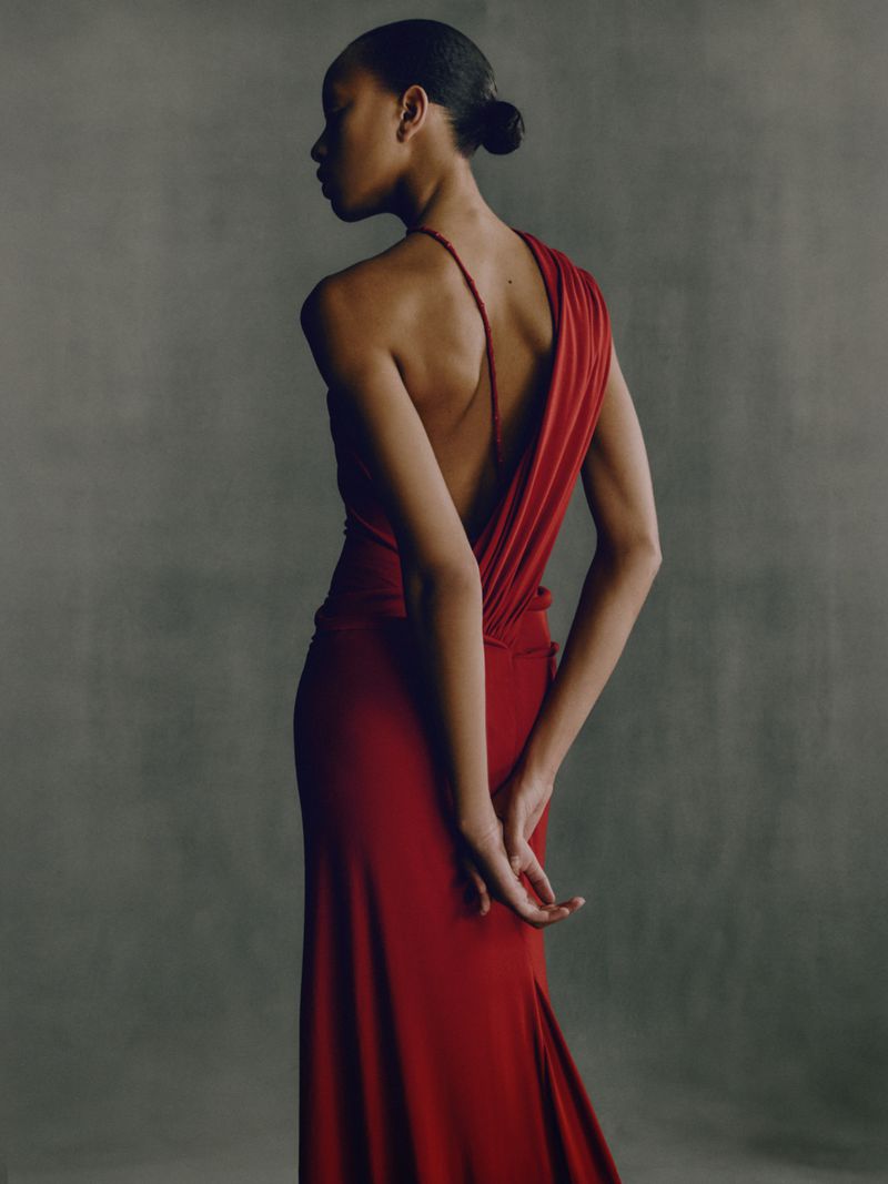 Elegant back view of an adult female in a long red gown, showcasing fashion and style against a neutral studio backdrop.