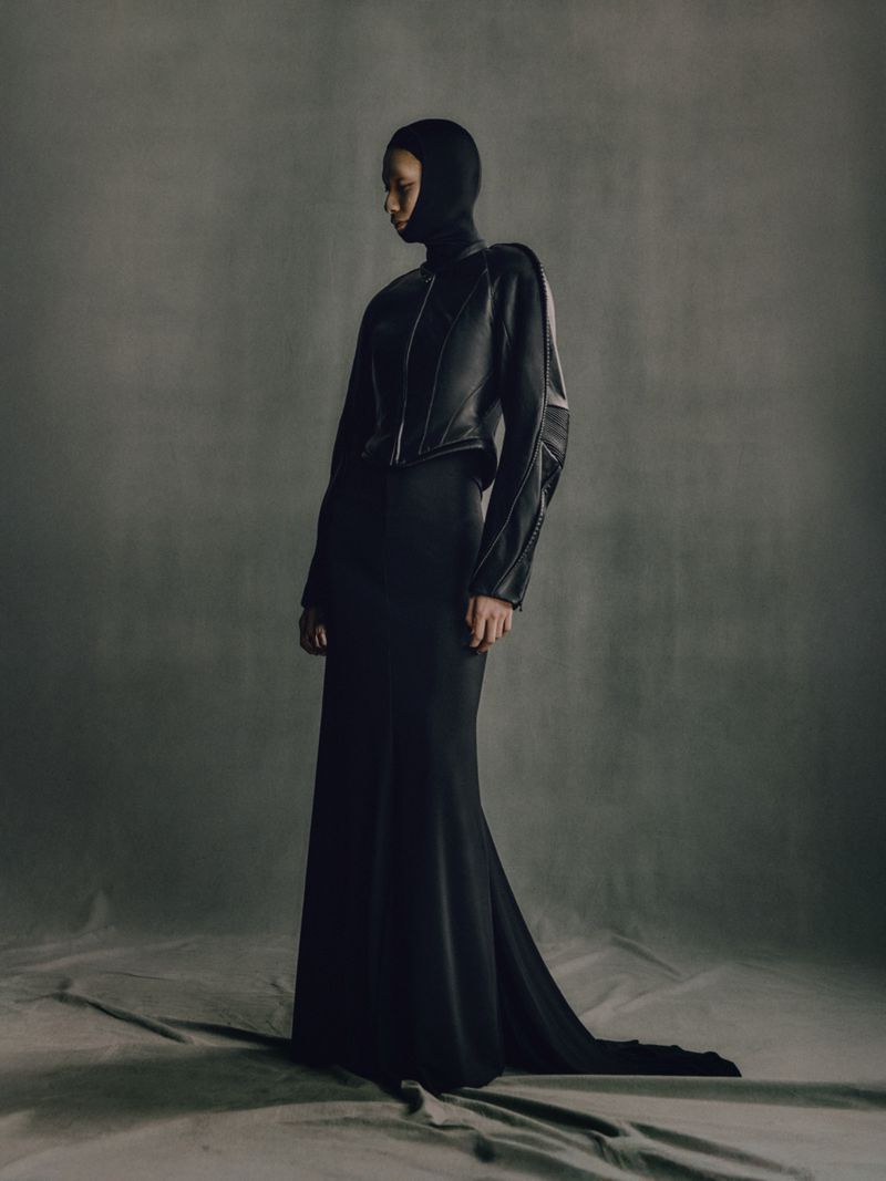 Elegant woman in a sleek, dark hooded dress and jacket poses against a neutral grey background in a fashion forward editorial shot.