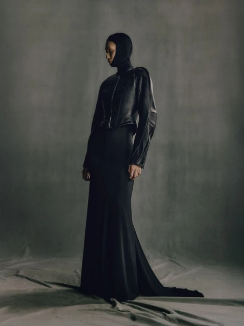 Elegant woman in a sleek, dark hooded dress and jacket poses against a neutral grey background in a fashion forward editorial shot.