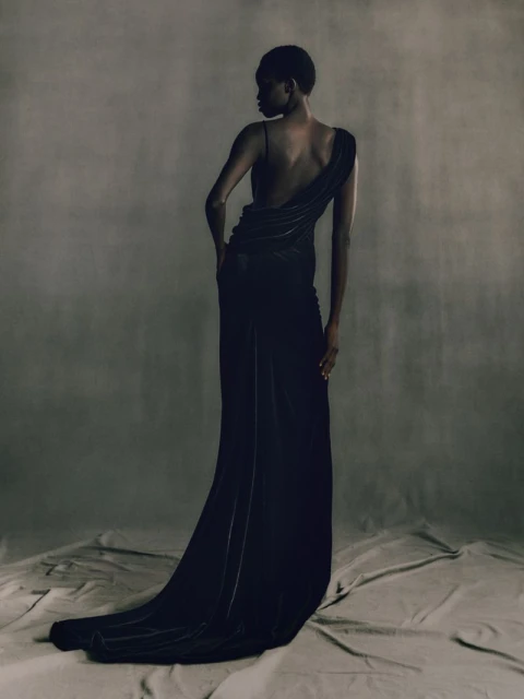 Elegant, slender Black female fashion model poses in a dark, flowing gown with one shoulder strap, against a textured grey background.