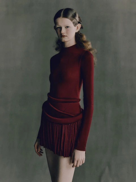 Woman with styled hair poses in a red long-sleeve top and matching fringed skirt against a grey background in a studio fashion shot.