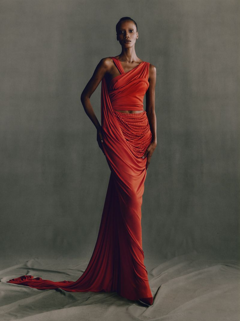 A slender woman models an orange draped gown in a studio setting, posing gracefully against a neutral backdrop with subtle floor textures.