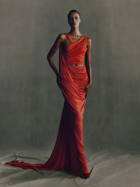 A slender woman models an orange draped gown in a studio setting, posing gracefully against a neutral backdrop with subtle floor textures.