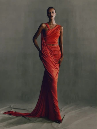 A slender woman models an orange draped gown in a studio setting, posing gracefully against a neutral backdrop with subtle floor textures.