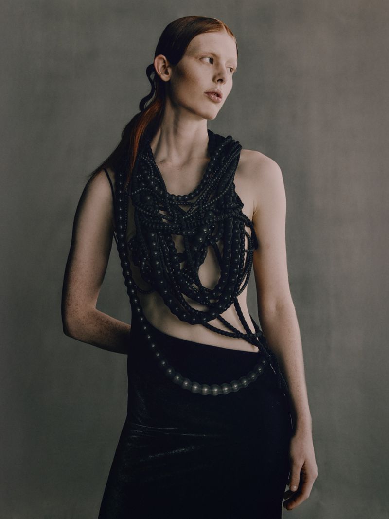 A slender woman with red hair poses in an elegant black gown and elaborate beaded collar against a neutral background, in a studio shot.