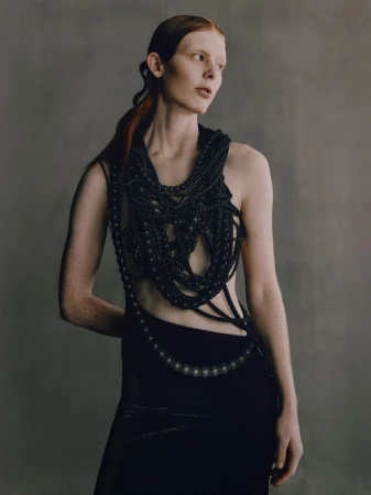 A slender woman with red hair poses in an elegant black gown and elaborate beaded collar against a neutral background, in a studio shot.