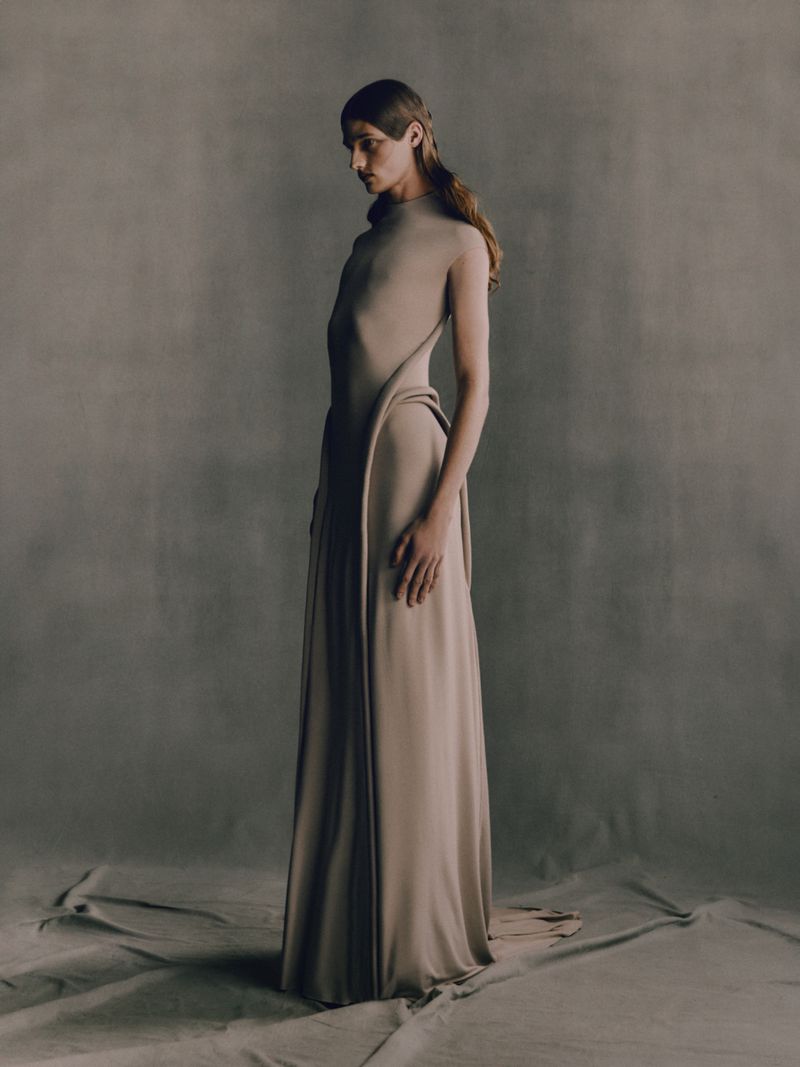 A slender figure in a beige gown stands in a minimalist studio setting, looking down with soft lighting that emphasizes the dress's draped design.