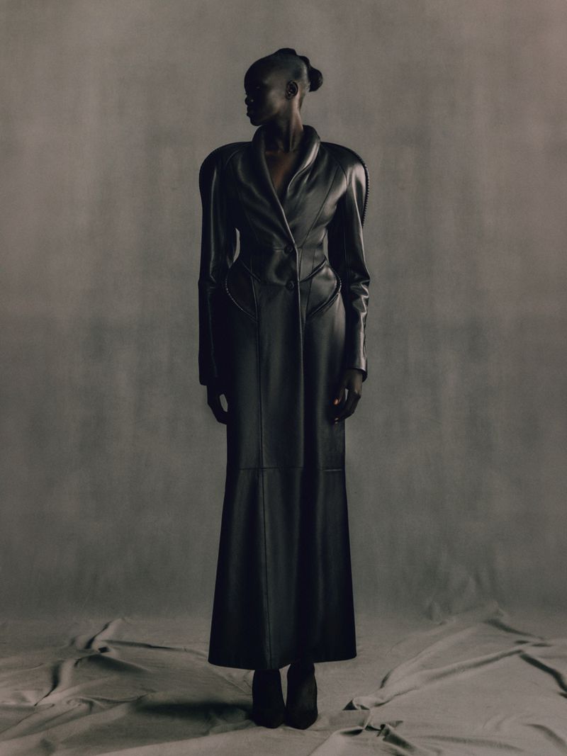 Full-length studio shot of an adult woman in a black leather trench coat, looking away from the camera, against a gray backdrop.