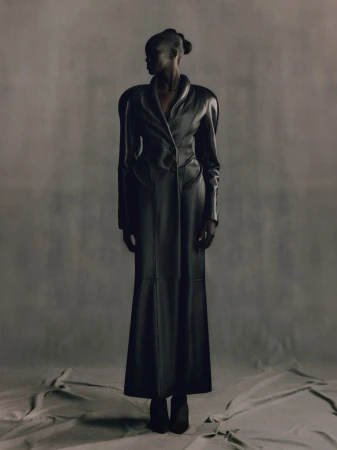 Full-length studio shot of an adult woman in a black leather trench coat, looking away from the camera, against a gray backdrop.