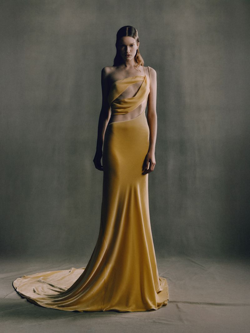 A woman stands in a yellow, floor-length formal dress with a train, posing against a grey backdrop in a fashion editorial style photograph.