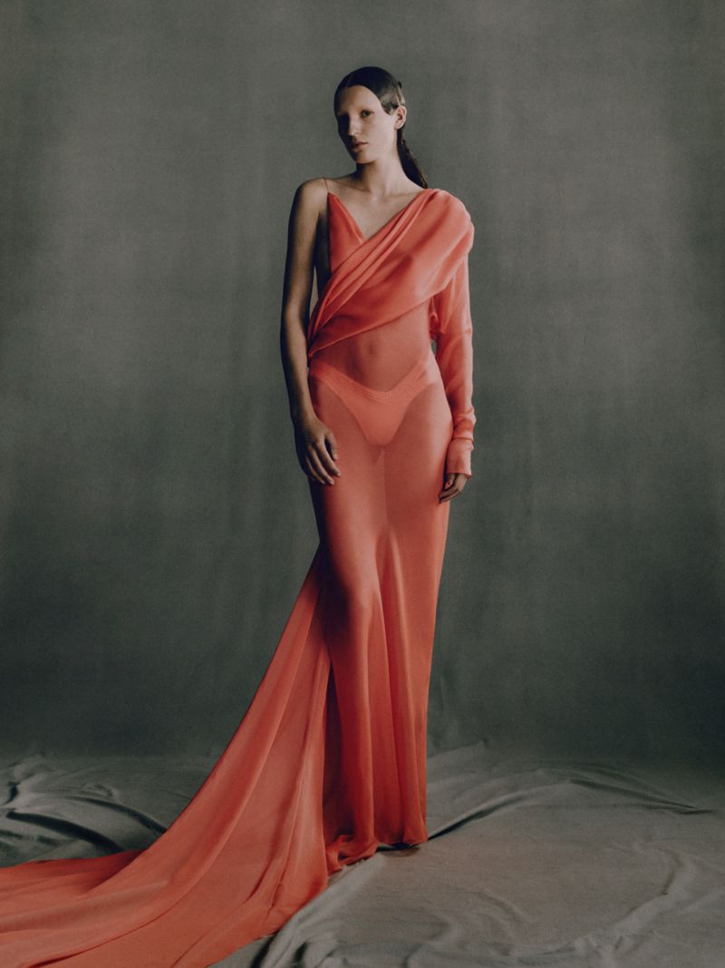 Elegant woman in a sheer coral gown with a train, posing in a neutral grey studio setting with soft lighting, capturing a high fashion moment.