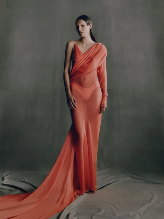 Elegant woman in a sheer coral gown with a train, posing in a neutral grey studio setting with soft lighting, capturing a high fashion moment.