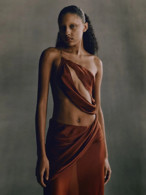 A woman poses in a draped, rust-colored dress against a soft grey background in a fashion photograph with a focus on texture and design.