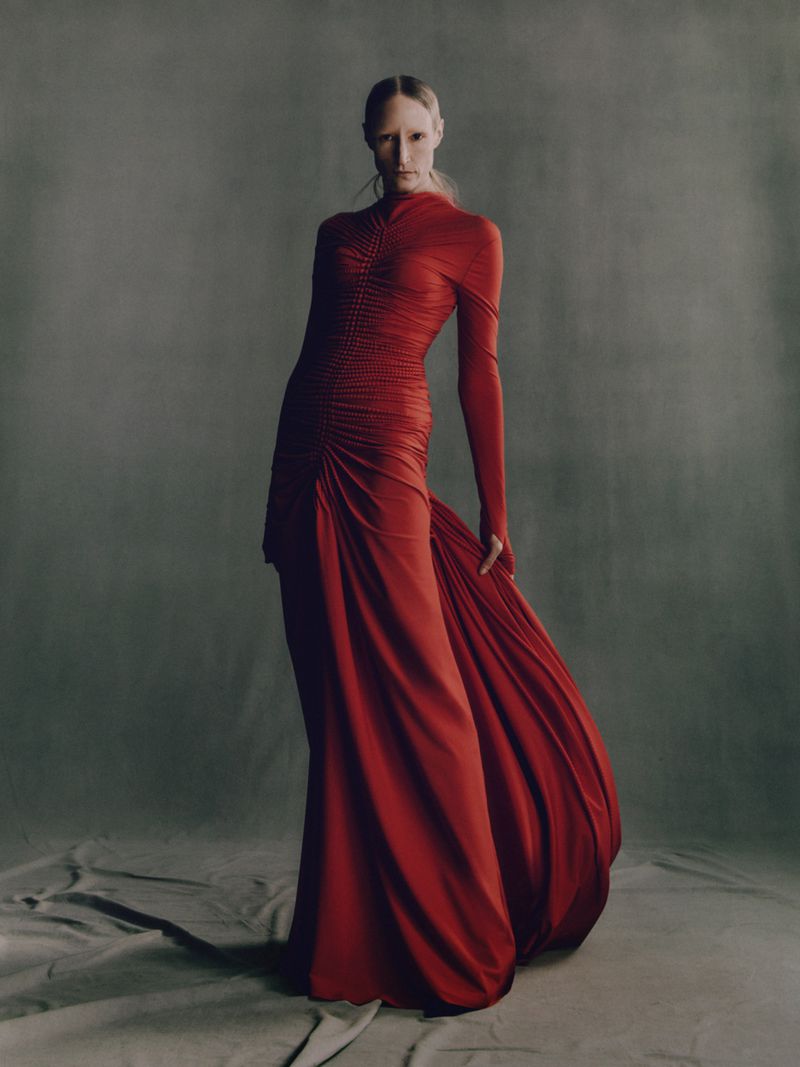 Elegant woman in long-sleeve red gown poses in a photo studio. Formal dress with train and gathered details against a grey backdrop.