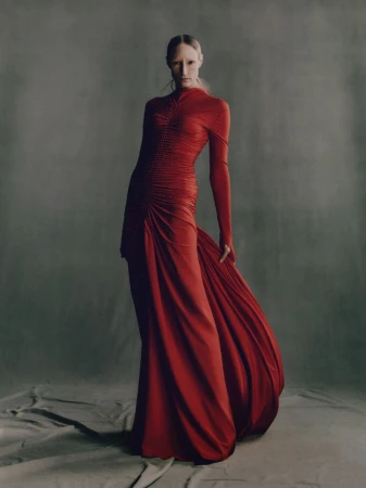Elegant woman in long-sleeve red gown poses in a photo studio. Formal dress with train and gathered details against a grey backdrop.