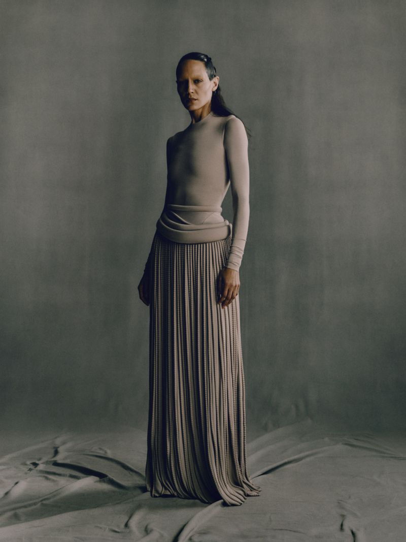 Woman in a long, flowing pleated skirt and long-sleeved top poses in a studio with a neutral background, showcasing a minimalist fashion style.
