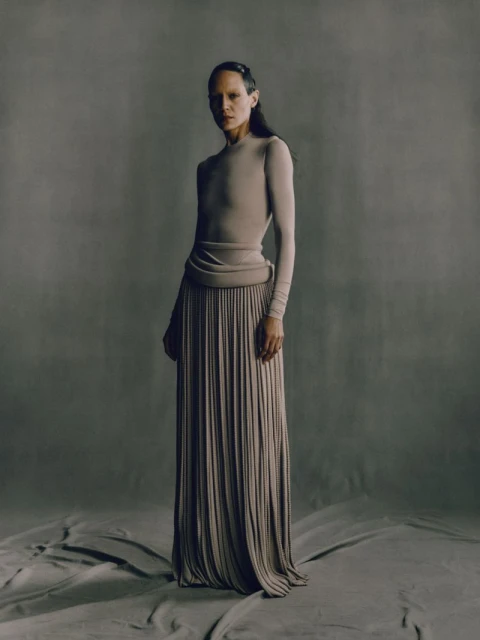 Woman in a long, flowing pleated skirt and long-sleeved top poses in a studio with a neutral background, showcasing a minimalist fashion style.