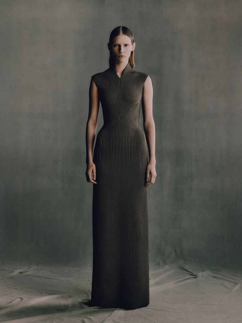 A full-length studio shot of a young adult woman in a sleeveless, dark green knit gown, posing against a mottled grey backdrop.