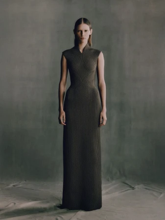 A full-length studio shot of a young adult woman in a sleeveless, dark green knit gown, posing against a mottled grey backdrop.