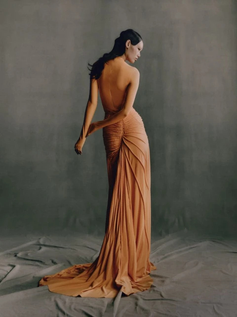 A woman in a peach-colored gown, seen from the back, poses in a photo studio with a draped grey backdrop and soft, diffused lighting.