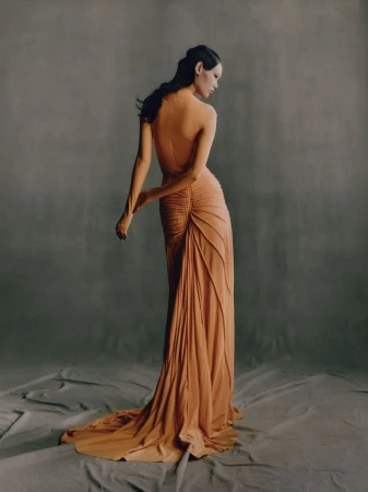 A woman in a peach-colored gown, seen from the back, poses in a photo studio with a draped grey backdrop and soft, diffused lighting.
