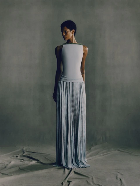 Woman in sleeveless top and long pleated skirt stands in a photo studio against a grey backdrop with soft, diffused lighting.