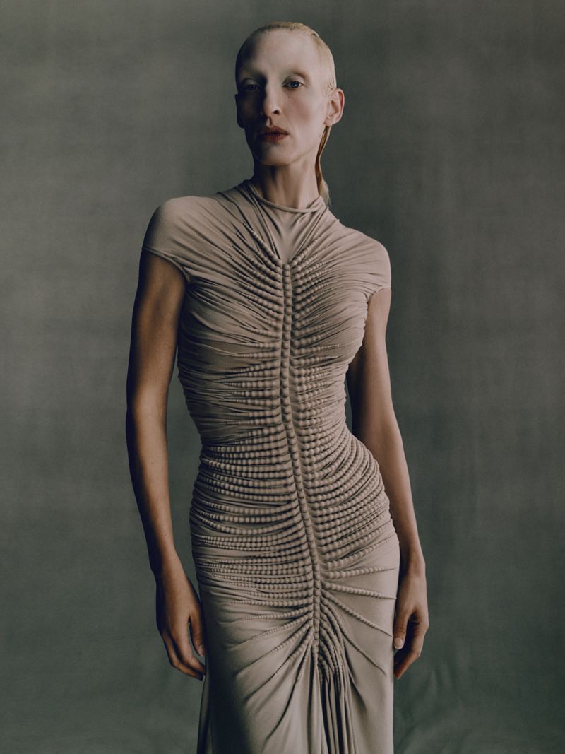 Adult female model with pale skin poses in a studio. She wears a beige form-fitting gown with gathering and beading along the torso.