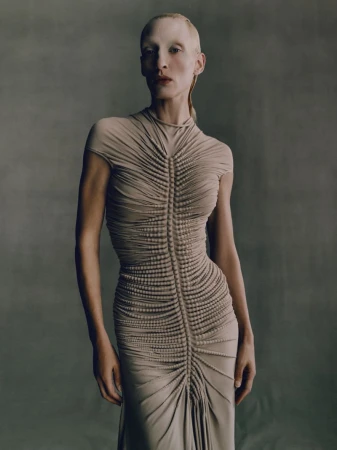 Adult female model with pale skin poses in a studio. She wears a beige form-fitting gown with gathering and beading along the torso.