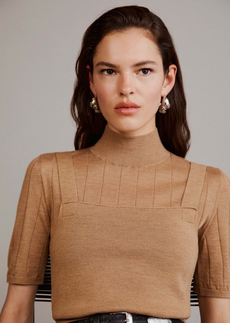 Brunette woman in a fitted brown sweater with a square neckline and silver hoop earrings, medium shot, neutral background, studio lighting.