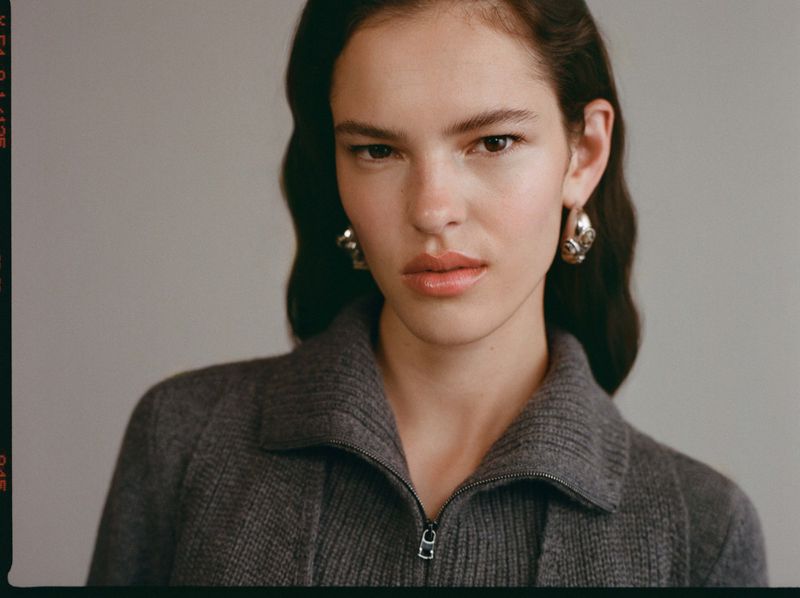Close-up studio shot of a young adult woman with wavy dark hair, wearing hoop earrings and a grey wool jacket. Neutral background.