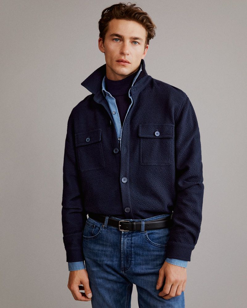 Man in navy jacket, blue turtleneck, and jeans stands in front of neutral backdrop in a medium studio shot. Fashion editorial.
