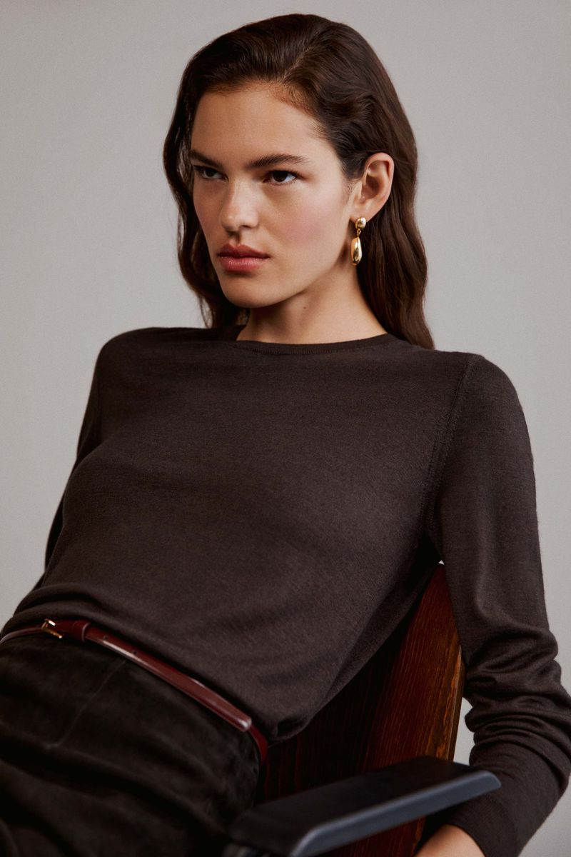 Adult woman wearing a long-sleeve brown shirt and dark pants sitting in a chair indoors with a plain gray background in a studio.
