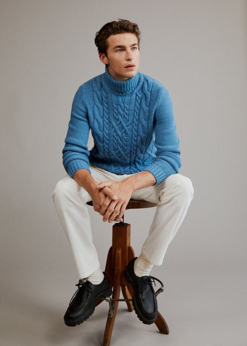 A young male model seated on a wooden stool wearing a blue cable-knit turtleneck, white trousers, and black shoes against a grey background.