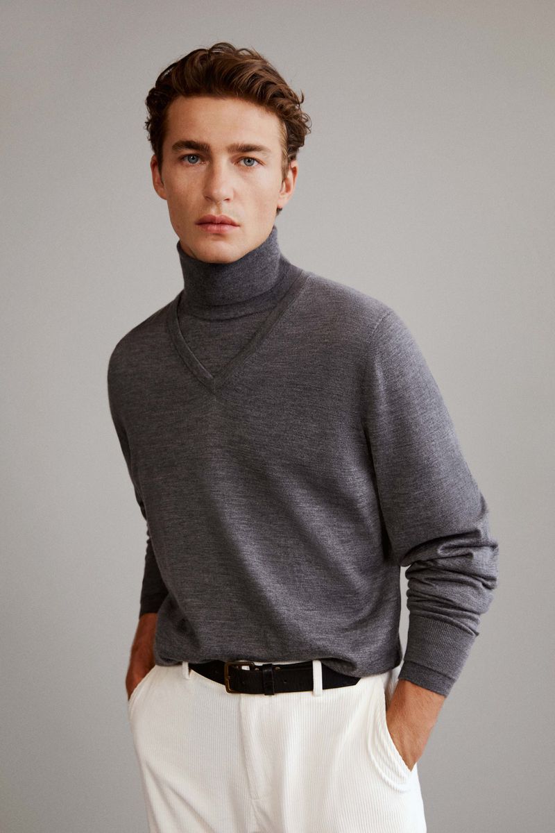 A handsome adult male poses in a studio wearing a gray turtle neck sweater, white pants, and a black belt against a gray backdrop.