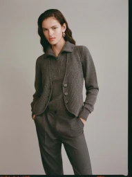 Model wearing a grey knitted cardigan and matching trousers against a solid neutral studio background.