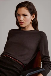 Model wearing dark knit top and belt sitting on a chair against a plain light grey studio background.
