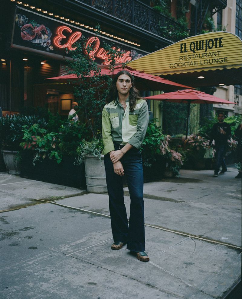 Person stands in front of El Quijote Restaurant, featuring a neon sign, plants, and yellow awning, on a city street.