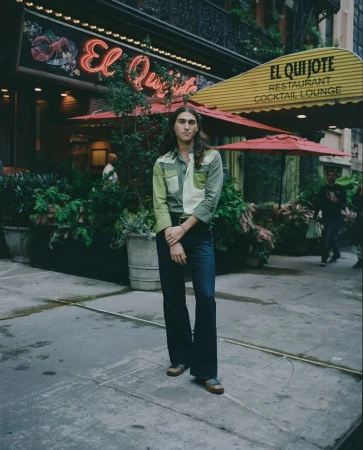 Person stands in front of El Quijote Restaurant, featuring a neon sign, plants, and yellow awning, on a city street.