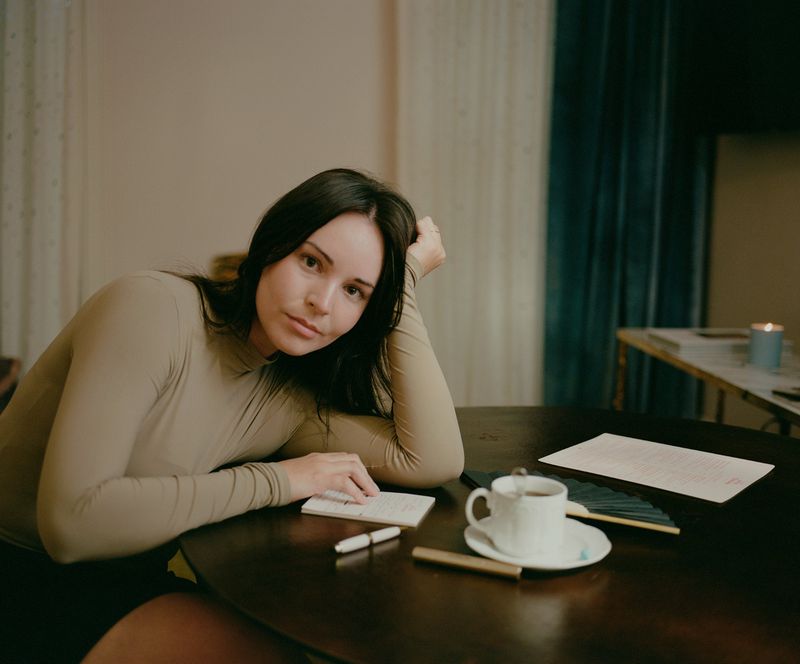 Woman with long dark hair leaning on a dark wooden table in an indoor room with a cup of coffee and notebook.
