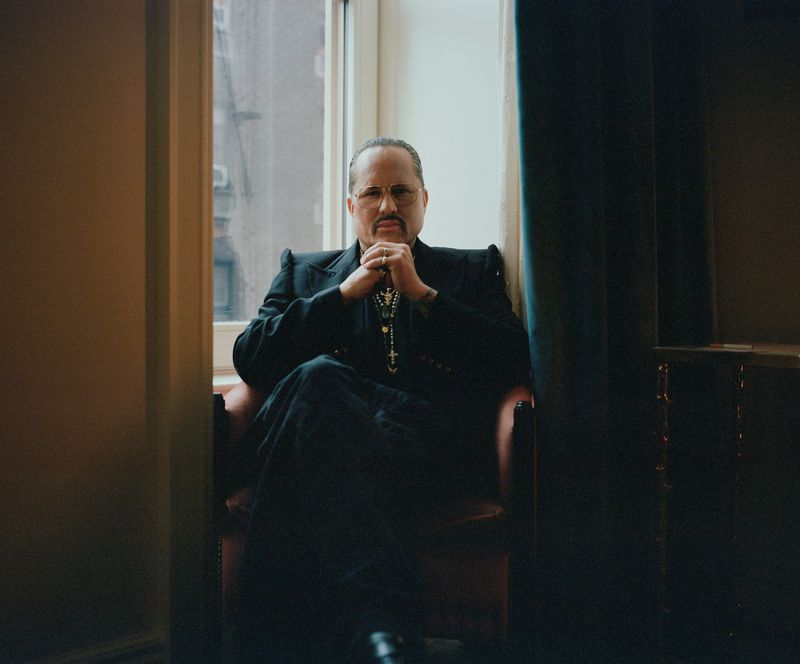 Portrait of an adult man with dark hair and a moustache, wearing a dark suit and silver cross necklace, seated by a window.