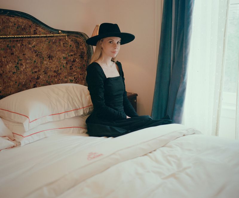 Woman wearing a black dress and black hat seated on a bed with white linens inside a hotel room.