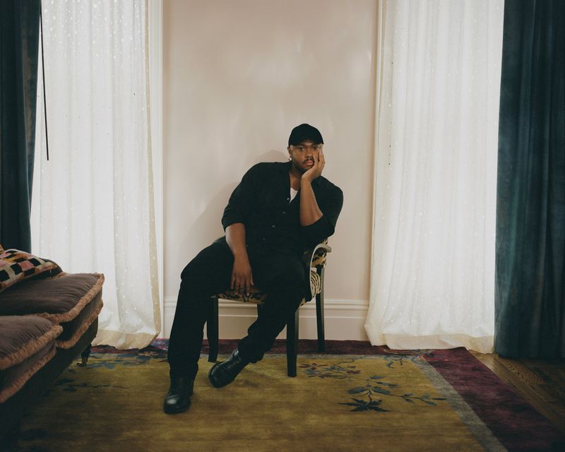A man in black casual attire sitting on a chair indoors in front of draped curtains, resting his head on his hand in a relaxed pose.