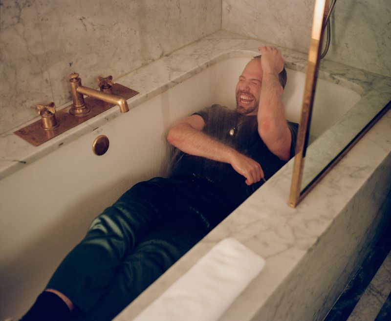 Man in bathtub laughing under running water, wearing dark shirt, surrounded by marble and gold fixtures in well-lit indoor bathroom.