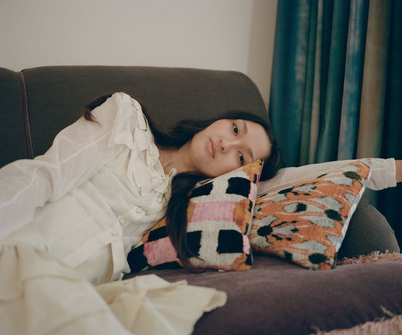 Adult woman in white dress rests on brown couch with patterned pillows, teal curtains and white wall in background in eye-level shot.