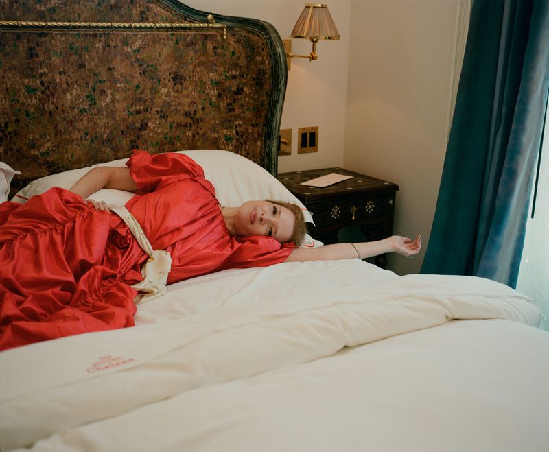 Woman in a red dress laying on a white bed in a hotel room with vintage decor, a nightstand, a lamp, and a blue curtain. 