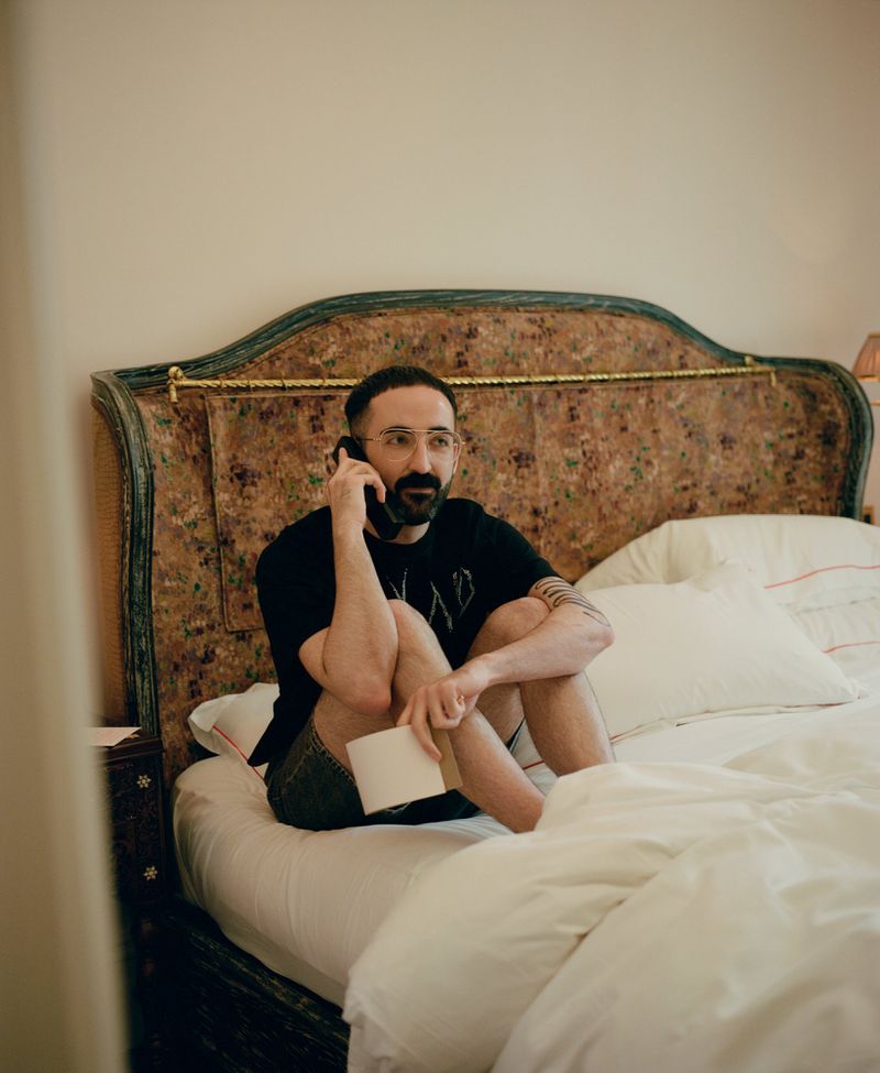 Adult man with a beard sits on a bed in a bedroom while on a phone call, holding a notepad, with a decorative headboard behind him.