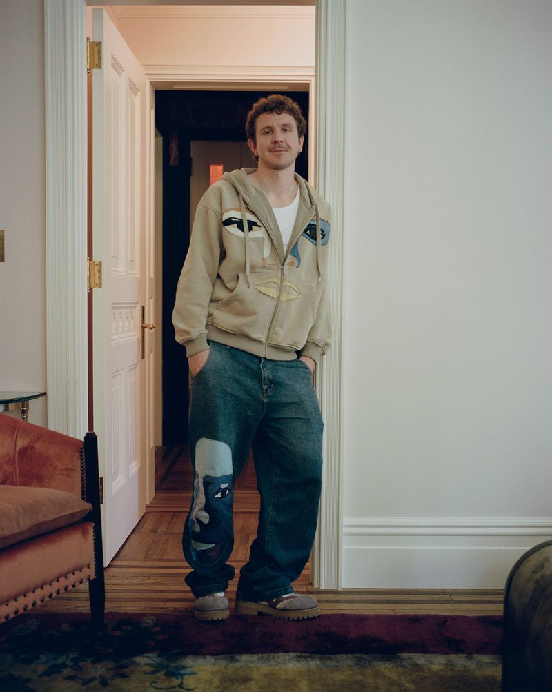 Indoor studio shot of an adult male in a beige hoodie and oversized jeans, standing in a doorway with a neutral facial expression.